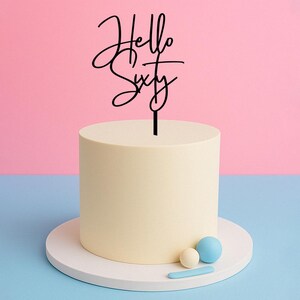 May include: Black acrylic cake topper with the text "Hello Sixty" in a cursive font. The topper is on top of a white cake with blue and white decorations.