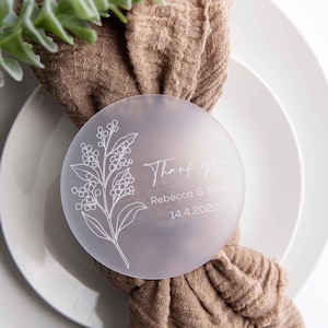 May include: A frosted glass round place card with a floral design and the text "Thank you, Rebecca & Kyle, 14.4.2022". The place card is tied to a brown fabric napkin.