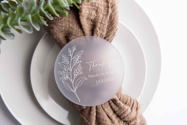 May include: A frosted glass round place card with a floral design and the text "Thank you, Rebecca & Kyle, 14.4.2022". The place card is tied to a brown fabric napkin.