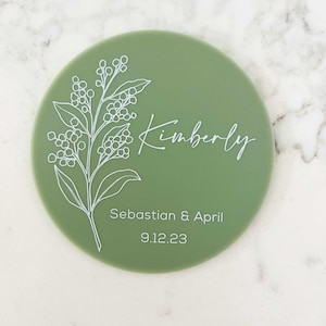 May include: A green round coaster with a white floral design and the text "Kimberly, Sebastian & April 9.12.23"