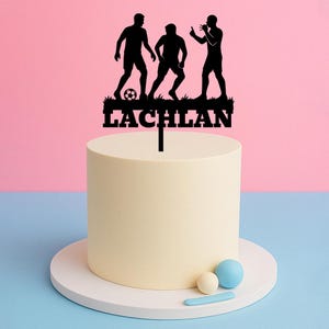 May include: Black silhouette cake topper of three soccer players with a soccer ball and the name Lachlan. The topper is on a white cake with a white cake stand.