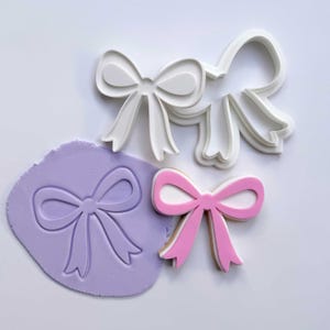 May include: A collection of bow-shaped cookie cutters and a decorated cookie. The cutters are white plastic, and the cookie is pink with a white center. A purple piece of dough has a bow imprint.