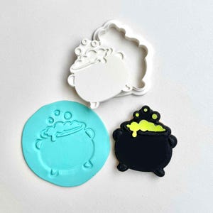 Puede incluir: Imagen de temática Halloween con un cortador de galletas de caldero negro, un molde turquesa y una galleta decorada. La galleta es negra con poción burbujeante amarilla. El cortador de galletas blanco tiene forma de caldero.