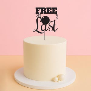 Op de afbeelding: Een zwarte taarttopper met de tekst "FREE at Last" en een kogel en ketting. De topper staat op een witte taart met witte parelversieringen.
