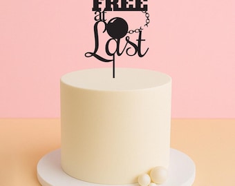 Puede incluir: Un adorno para pastel negro con el texto "FREE at Last" y una bola y una cadena. El adorno está sobre un pastel blanco con decoraciones de perlas blancas.