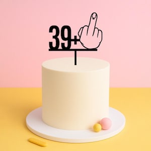 May include: A white cake with a black cake topper that says "39+" with a middle finger. The cake topper is on a white cake stand.