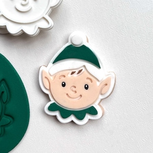 May include: A cookie cutter in the shape of an elf's face. The elf has a green hat and collar, with a white trim. The face is light brown with black eyes and a smiling mouth. The cookie cutter is white.