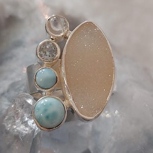 Könnte beinhalten: Ein silberner Ring mit einem großen, ovalen, beigefarbenen Druzy-Stein und drei kleineren, runden, hellblauen und klaren Steinen. Der Ring hat ein einzigartiges, asymmetrisches Design, ideal für einen Hauch von Eleganz.