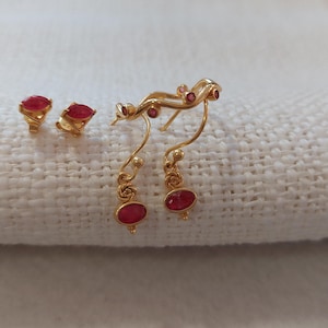 Op de afbeelding: Een paar gouden oorbellen met rode edelsteenaccenten. De oorbellen hebben een hangend ontwerp met een gebogen, rankvorm.