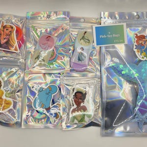 May include: A collection of seven holographic resealable bags containing various character-themed stickers and accessories. The bags feature images of animated characters, including a mermaid, a princess, and a genie, with a Pick-See Bags ETS-54 label.