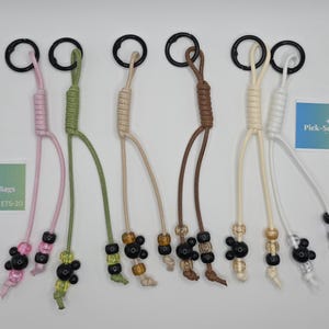 May include: Six bag charms featuring colored cords attached to black metal rings. Each charm has knotted cord sections and decorative beads, including black spherical beads. Two small rectangular cards with the text Pick-See Bags ETS-20 are present.