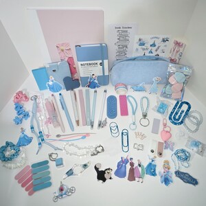 May include: A collection of stationery and accessories featuring a light blue and pink color palette, including a notebook, pencil case, pens, stickers of Cinderella characters, hair scrunchies, paper clips, keychains, and various small decorative items.