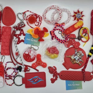 May include: A collection of red-themed sensory toys and accessories including lanyards, fidget spinners, pop-it keychains, silicone textured strips, beaded bracelets, and a circular disc featuring a cartoon character graphic with Pick-See Bags text.