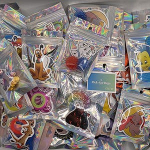 May include: A collection of small, holographic, resealable plastic bags, each containing a unique character sticker and a small accessory. A central white rectangular card features the text Pick-See Bags and ETS-57. The stickers depict various animated characters, including a yellow dog, a blue fish, a gray elephant, and a purple cat.