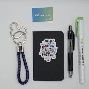 May include: A black notebook with a sticker of cartoon characters, a blue and black braided keychain with a Mickey-shaped ring, a black retractable ballpoint pen, a light green highlighter, and a small rectangular card labeled Pick-See Bags ETS-22.