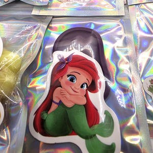 May include: A die-cut sticker featuring a cartoon mermaid with long red hair, a purple hair accessory, and a green tail, shown in a seated pose with arms crossed. The sticker is packaged inside a clear, iridescent, holographic plastic pouch.