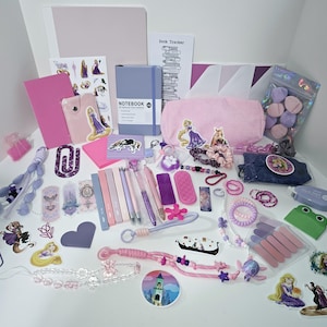 May include: A collection of stationery and accessories in shades of pink and purple, including a notebook, a pencil case, various pens, stickers featuring a long-haired character, bookmarks, hair ties, a beaded lanyard, and a small green frog-shaped item.