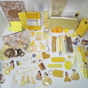 May include: A collection of yellow-themed stationery and accessories including a notebook, pencil case, pens, stickers featuring Belle and the Beast, hair scrunchies, keychains, paper clips, and various decorative items arranged on a white surface.