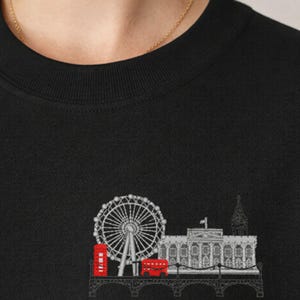 May include: Black sweatshirt featuring an embroidered London skyline design. The design includes the London Eye, a red telephone booth, a double-decker bus, and other iconic landmarks. The sweatshirt has a crew neck and long sleeves.