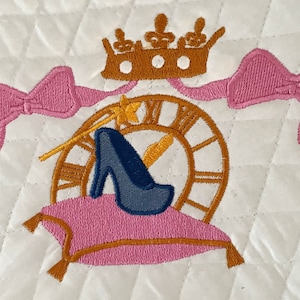 May include: Embroidered design featuring a blue glass slipper, a gold clock, a gold crown, and pink bows on a white quilted fabric.