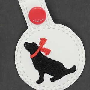 May include: White leather key chain with a red button and a black embroidered dog wearing a red scarf.