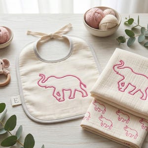 May include: A baby gift set featuring a cream-colored bib with a pink elephant embroidery, a matching burp cloth, and a crochet elephant rattle. The set includes a small bowl with pink yarn and a wooden letter "E".