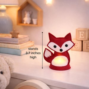 May include: A red felt fox-shaped night light, 3.7 inches high, with white accents and a warm glow. The fox has black eyes and a white belly. The text on the image says "stands 3.7 inches high".