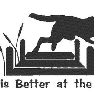May include: Black embroidered design featuring a dog leaping over a wooden dock, with cattails on the left. The text "Life is Better at the Lake" is below the image. The design is on a white background.