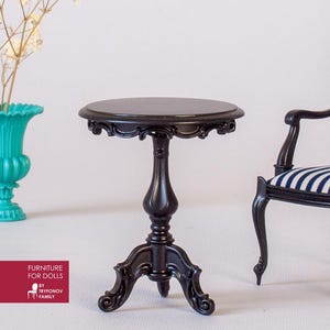 Puede incluir: Una mesa auxiliar negra en miniatura con detalles ornamentados, con una tapa redonda y una base de pedestal. La mesa es parte de un juego de muebles para casas de muñecas. Un jarrón turquesa con flores blancas y una silla a rayas están en el fondo.