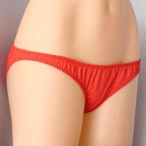 May include: Bright red underwear with a delicate, ruffled waistband. The brief-style bottoms are made of a soft, smooth fabric. The underwear is displayed on a mannequin.