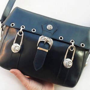 May include: Black leather crossbody bag with silver-tone hardware. Features a buckle closure, decorative rivets, and two large safety pin charms with lion head details. The bag has a shoulder strap.