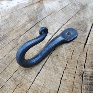 May include: A black metal hook with a decorative swirl at the top. The hook is mounted on a wooden surface.