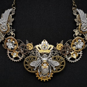 May include: A silver and gold necklace featuring a bee design at its centre. The bee is wearing a crown and is surrounded by gears and flowers. The necklace is made of metal and has a steampunk aesthetic.
