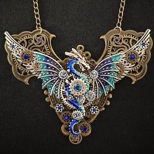 May include: A blue and green dragon with wings made of gears and rhinestones, set in a bronze frame. The dragon is part of a necklace with a chain.