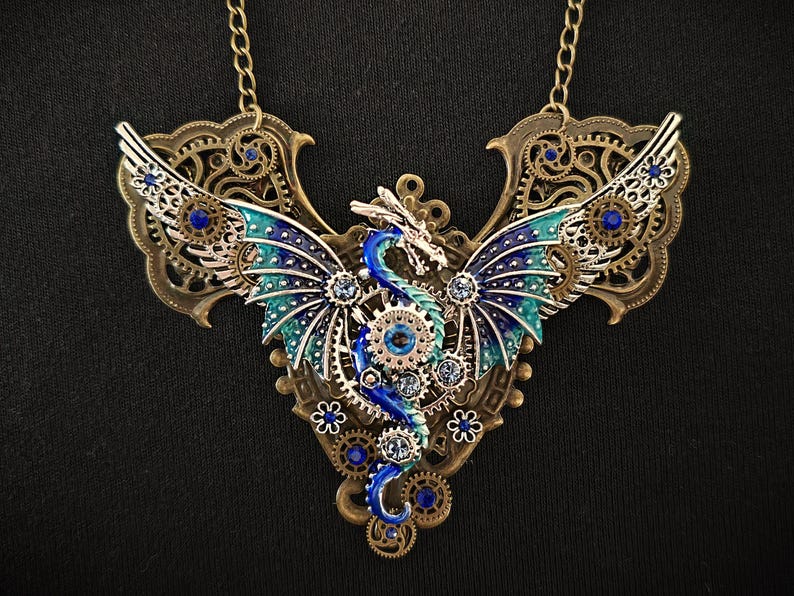 May include: A blue and green dragon with wings made of gears and rhinestones, set in a bronze frame. The dragon is part of a necklace with a chain.