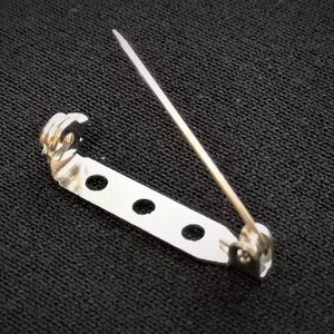 May include: Silver-toned metal safety pin with a decorative loop at the top and four holes along the bar. The pin is designed for attaching fabric or other materials.