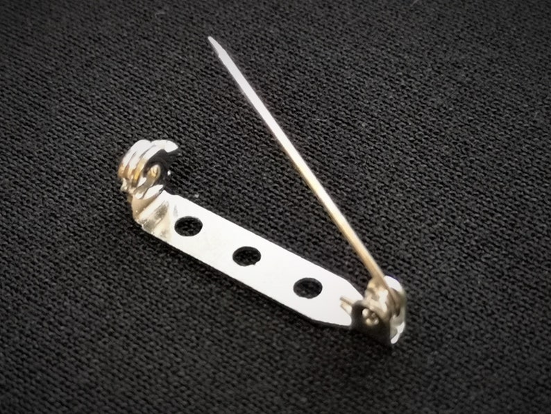 May include: Silver-toned metal safety pin with a decorative loop at the top and four holes along the bar. The pin is designed for attaching fabric or other materials.