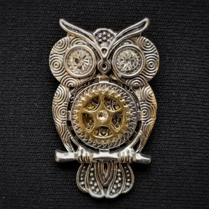 May include: A silver and gold owl pendant with a detailed design and a gear in the center. The owl's eyes are adorned with clear rhinestones.