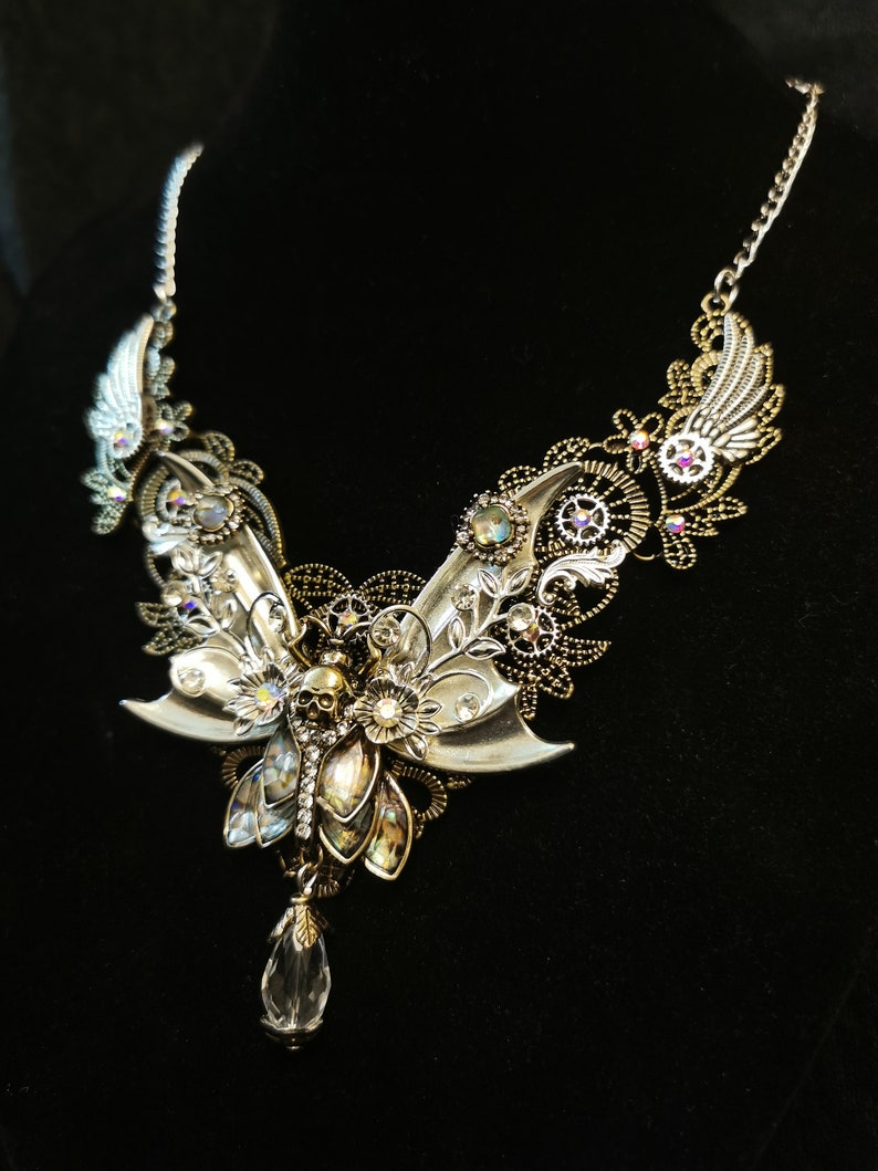 K&ouml;nnte beinhalten: Eine silberne und goldene Halskette mit einem gro&szlig;en, detaillierten Schmetterlingsanh&auml;nger. Der Schmetterling hat einen Sch&auml;del in der Mitte und ist von Kristallen und Zahnr&auml;dern umgeben. Die Halskette befindet sich auf einem schwarzen Hintergrund.