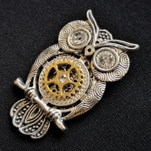 May include: A silver owl pendant with a gold gear design and clear rhinestones. The owl's wings are detailed with intricate patterns.
