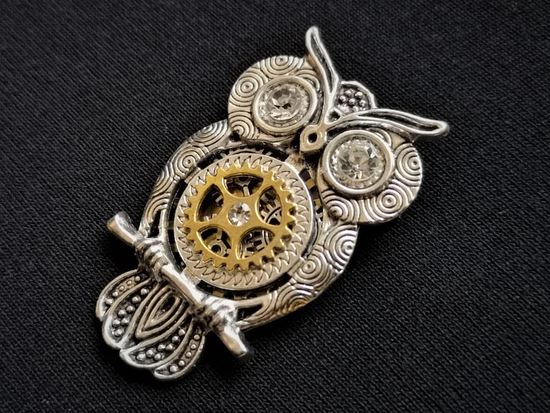 May include: A silver owl pendant with a gold gear design and clear rhinestones. The owl's wings are detailed with intricate patterns.
