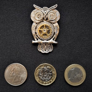 May include: A silver and gold owl brooch with a gear design. The owl is perched on a branch and has a detailed design. The brooch is surrounded by three coins: a US quarter, a British one pound coin, and a Euro coin.