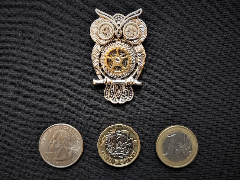 May include: A silver and gold owl brooch with a gear design. The owl is perched on a branch and has a detailed design. The brooch is surrounded by three coins: a US quarter, a British one pound coin, and a Euro coin.