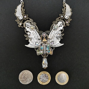 K&ouml;nnte beinhalten: Eine Halskette aus Silber und Bronze mit einem Totenkopf- und Schmetterlingsdesign. Die Schmetterlingsfl&uuml;gel sind aus irisierendem Abalone-Muschel gefertigt und die Halskette ist mit Kristallen verziert. Die Halskette wird auf einem schwarzen Hintergrund mit drei M&uuml;nzen zur Gr&ouml;&szlig;enbestimmung gezeigt.