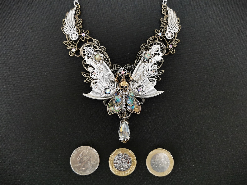 K&ouml;nnte beinhalten: Eine Halskette aus Silber und Bronze mit einem Totenkopf- und Schmetterlingsdesign. Die Schmetterlingsfl&uuml;gel sind aus irisierendem Abalone-Muschel gefertigt und die Halskette ist mit Kristallen verziert. Die Halskette wird auf einem schwarzen Hintergrund mit drei M&uuml;nzen zur Gr&ouml;&szlig;enbestimmung gezeigt.