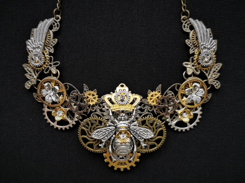 May include: A silver and gold necklace featuring a bee design at its centre. The bee is wearing a crown and is surrounded by gears and flowers. The necklace is made of metal and has a steampunk aesthetic.