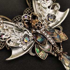 K&ouml;nnte beinhalten: Eine silberne und goldene Schmetterlingskette mit einem Totenkopf und Abalone-Muschel-Akzenten. Die Schmetterlingsfl&uuml;gel sind mit floralen Designs und Strasssteinen verziert.