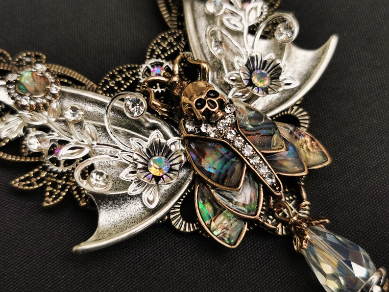 K&ouml;nnte beinhalten: Eine silberne und goldene Schmetterlingskette mit einem Totenkopf und Abalone-Muschel-Akzenten. Die Schmetterlingsfl&uuml;gel sind mit floralen Designs und Strasssteinen verziert.