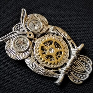 May include: A silver and gold owl pendant with intricate details and clear rhinestones. The owl's eyes are set with rhinestones and the body features a detailed gear design.