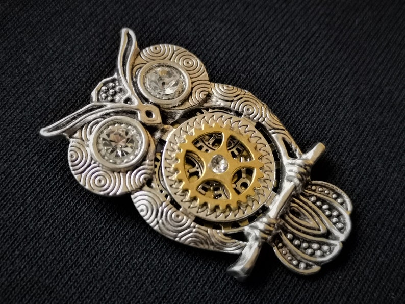 May include: A silver and gold owl pendant with intricate details and clear rhinestones. The owl's eyes are set with rhinestones and the body features a detailed gear design.